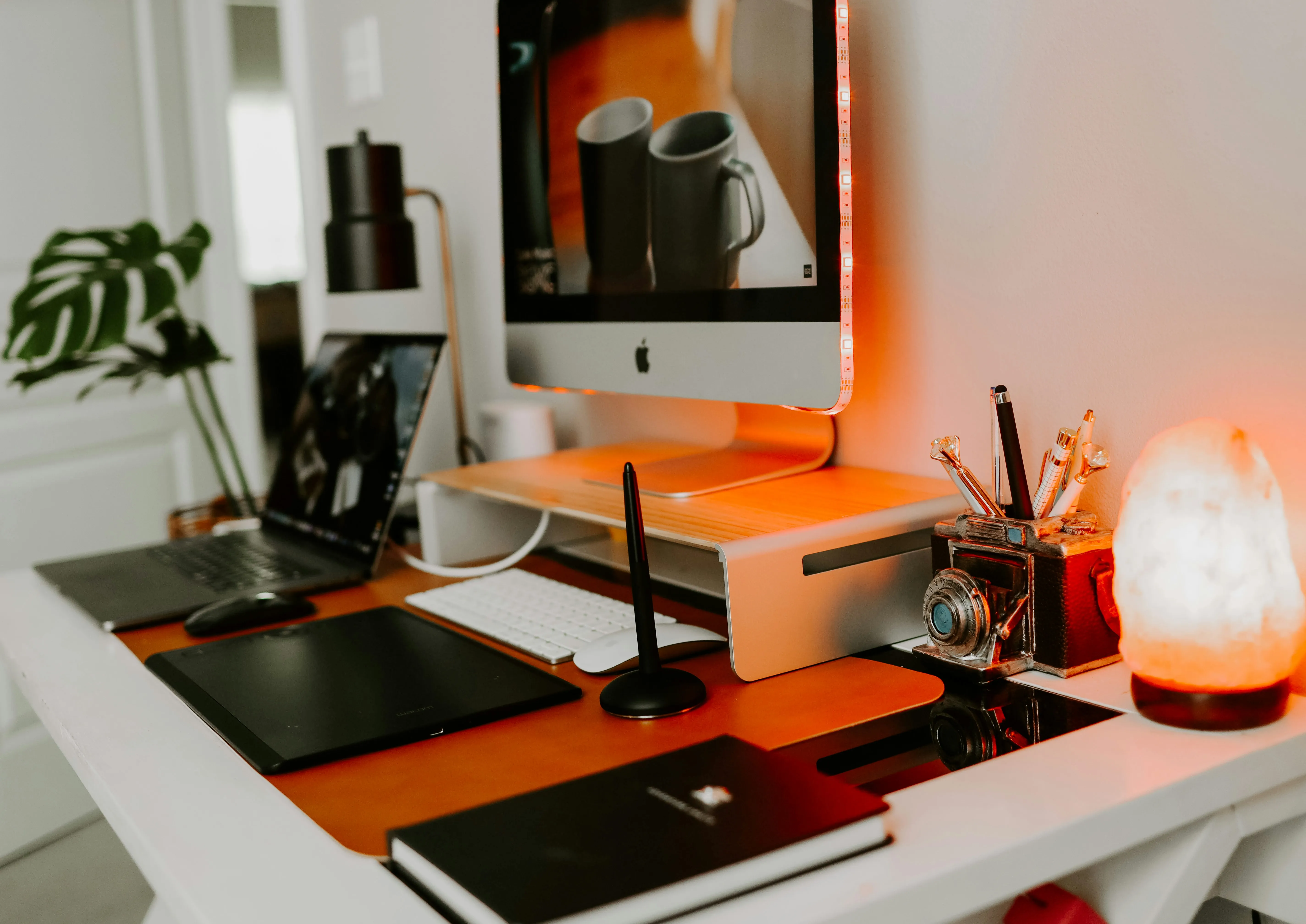Home office desk with an iMac on a monitor stand, laptop, drawing tablet, and salt lamp