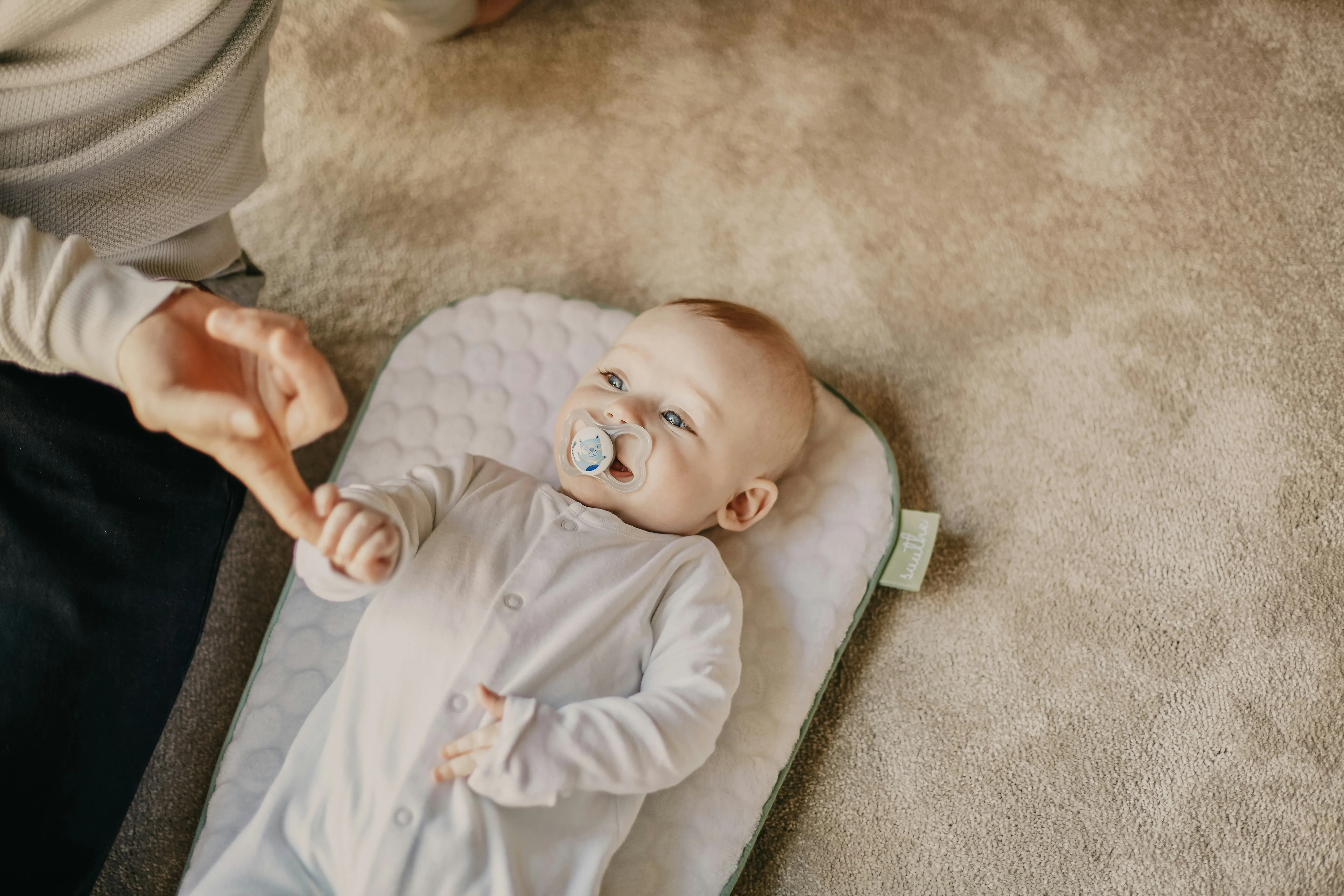Baby with a pacifier lying on a changing mat, holding an adult's finger