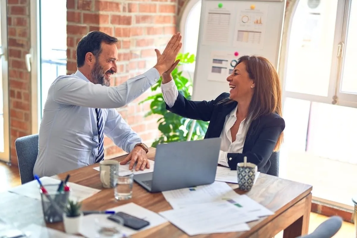 Two colleagues sharing a high five at a desk with a laptop and papers