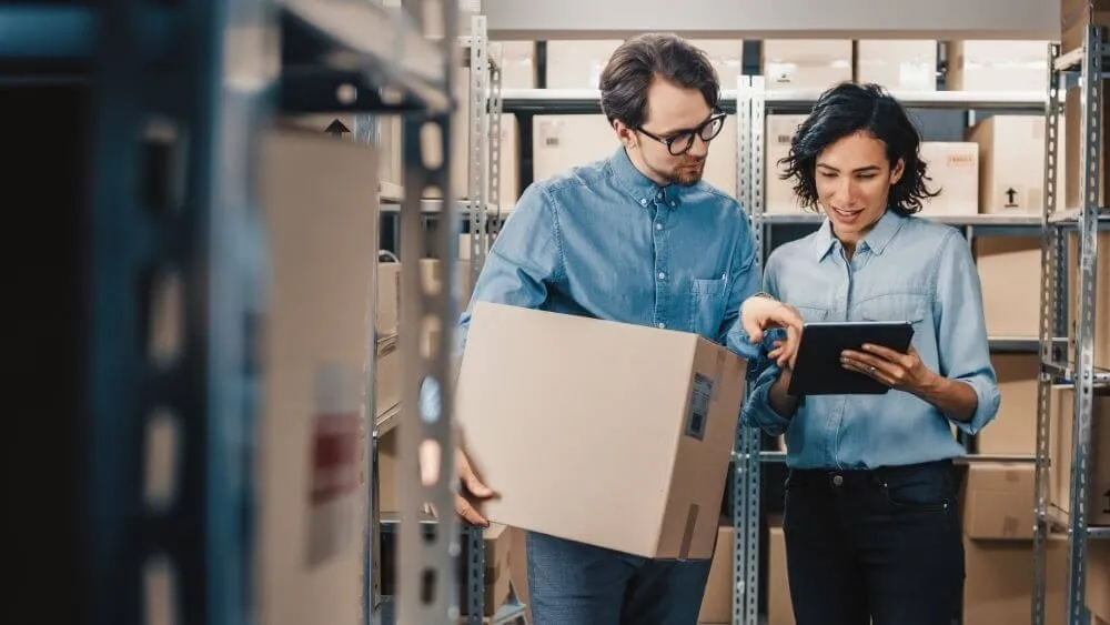 Two warehouse workers between stocked shelves, one carrying a box and one holding a tablet