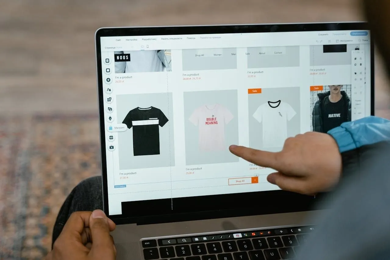 Person pointing at a laptop showing an online store's T-shirt product grid