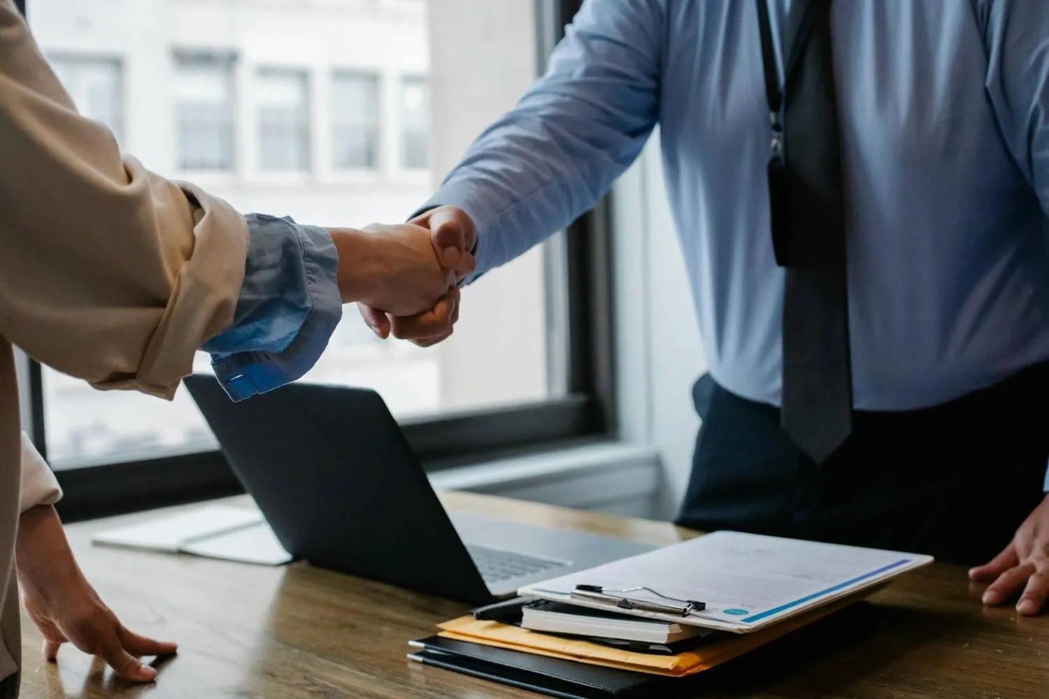 Two people shaking hands over a desk with a laptop and clipboards