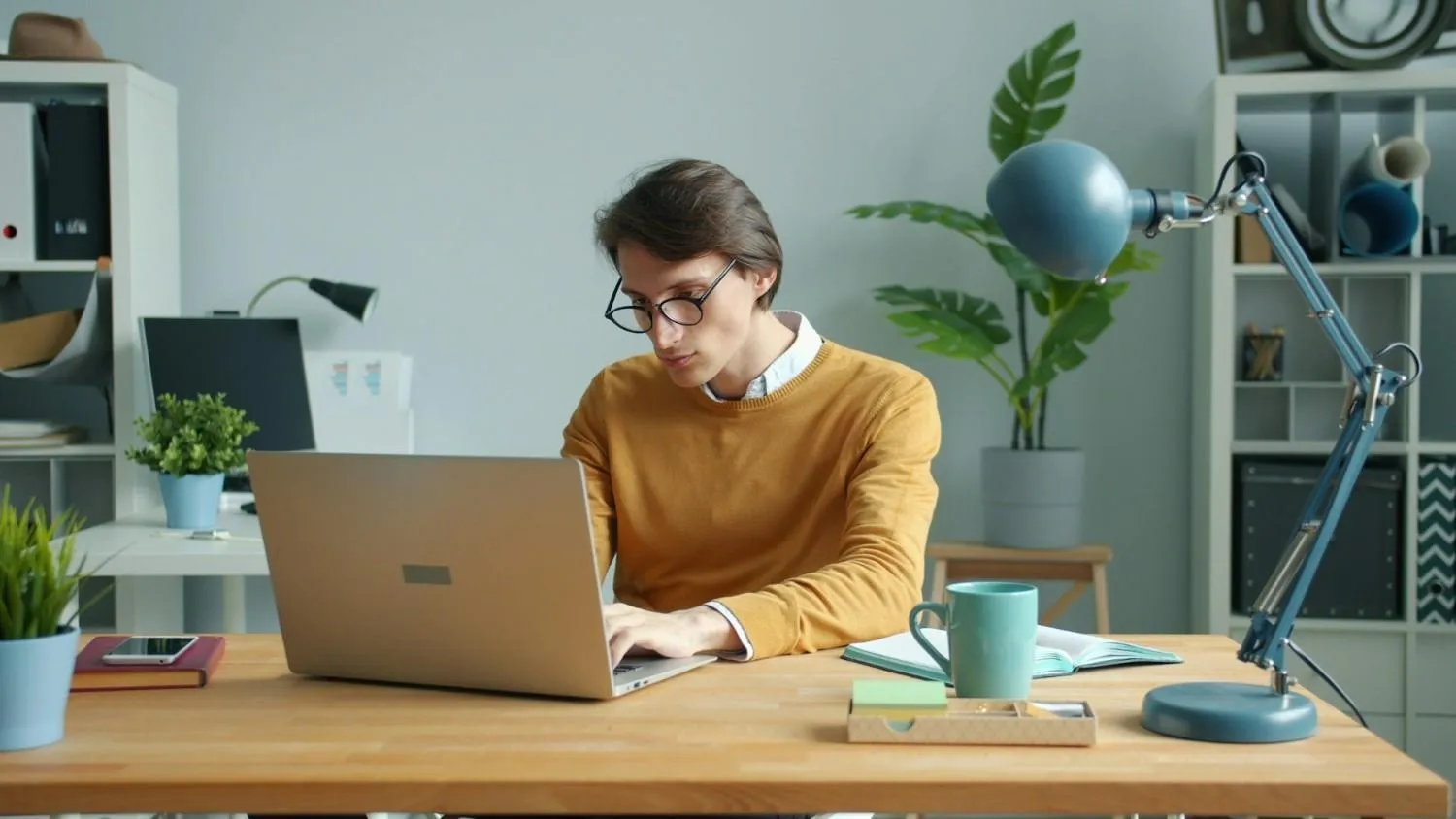 Man in a mustard sweater typing on a laptop at a home office desk
