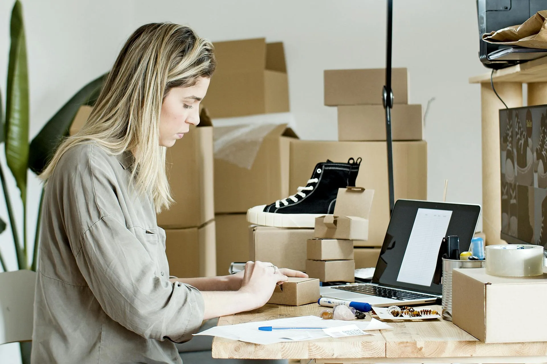 Woman packing a small box at a desk surrounded by shipping boxes and a sneaker