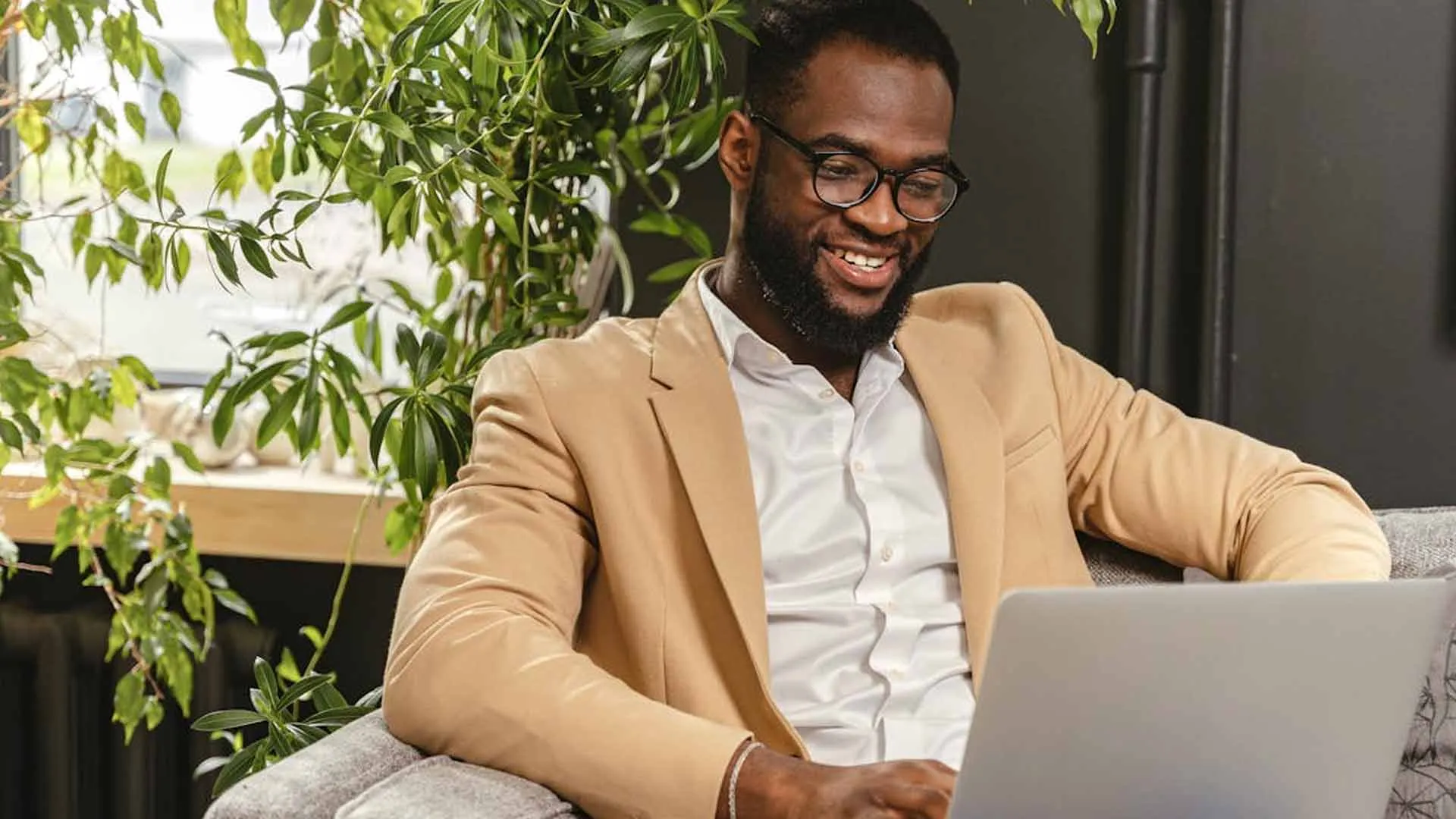 Smiling man in a tan blazer working on a laptop beside a plant