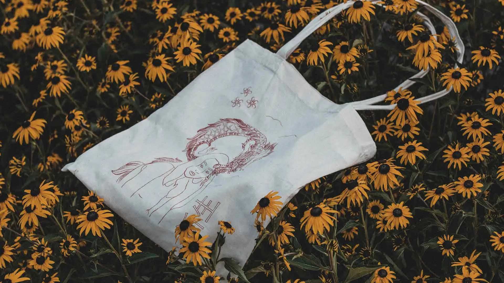 White canvas tote bag with a red line drawing of a dragon, lying among yellow flowers